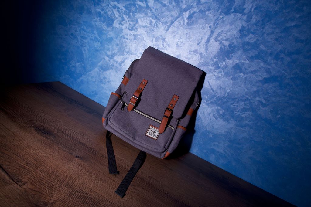 Photo by Salim Da- A modern backpack with leather straps sits on a wooden surface against a textured blue wall

